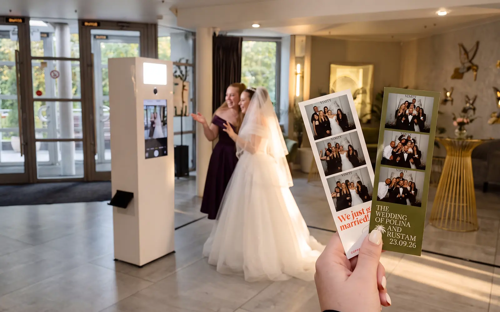 Невеста с подругой позируют перед белой фотобудкой в холле ресторана Vinity, в руке свадебные фотополоски 5×15